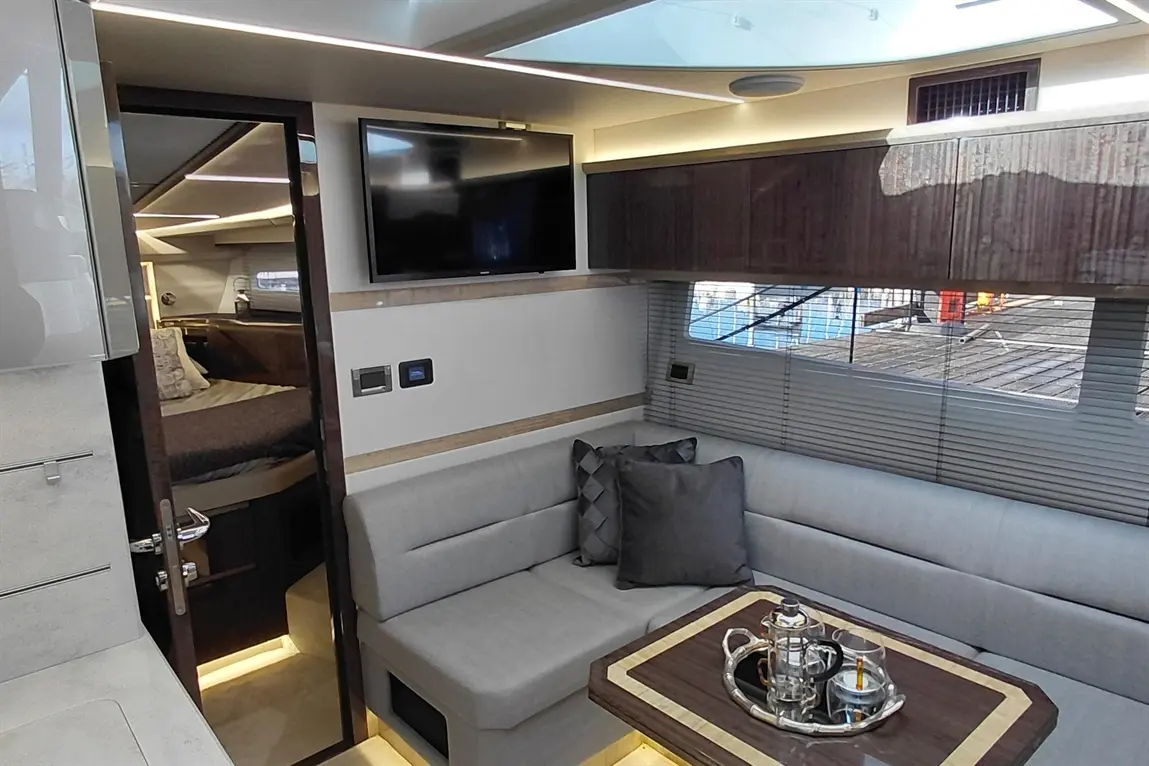 Modern yacht interior featuring a lounge area with grey sofa, coffee table, TV, and doorway leading to a bedroom.